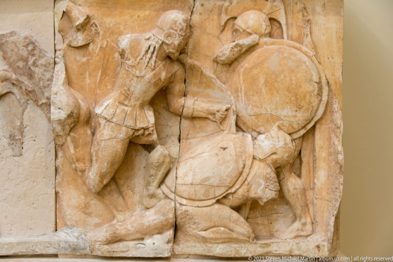 Fragmentary marble relief of a kneeling warrior crouched behind a large round shield.
