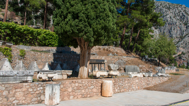 Rows of ancient column drums, capitals, and carved stone blocks displayed beside a path under trees at Delphi.