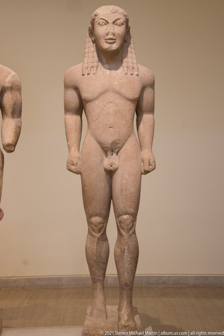 Front view of an Archaic male kouros with long patterned hair, clenched fists, and left leg advanced.