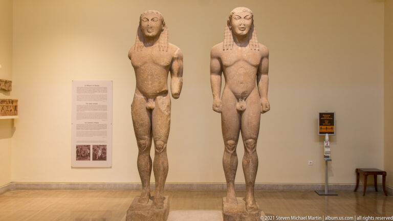 Two monumental Archaic male kouroi standing side by side in the Delphi Archaeological Museum.