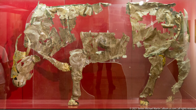 Fragmentary gold sheets and pale ivory body parts from an ancient composite figure displayed against red backing.