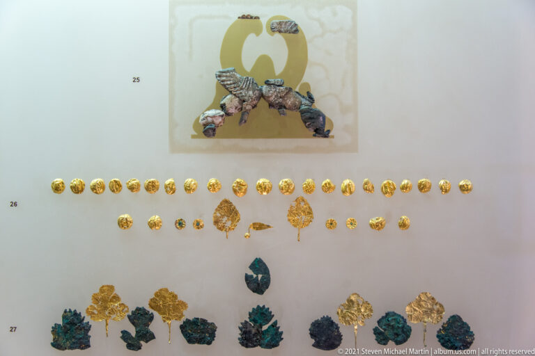 Museum display of rows of small gold disks, leaf-shaped ornaments, and darker decorative pieces.