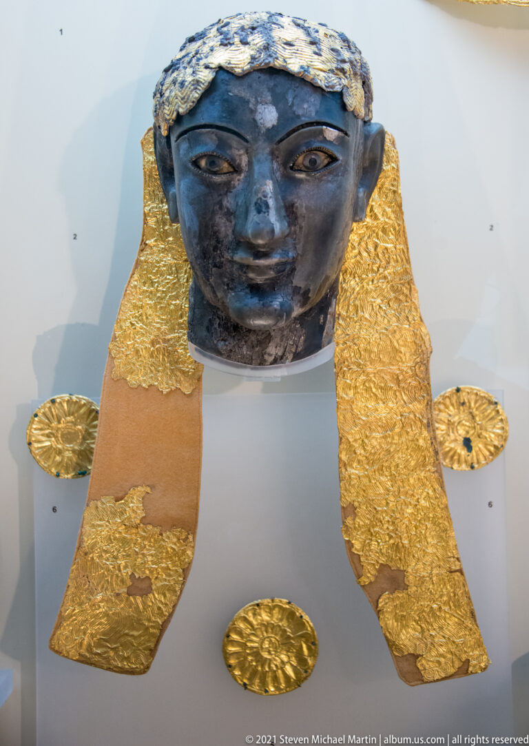 Dark ancient female head displayed with broad hammered-gold headdress or hair elements and separate gold appliqués.