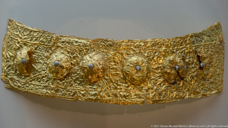 Curved hammered-gold band decorated with raised rosettes, dark central studs, and dense repoussé patterns.