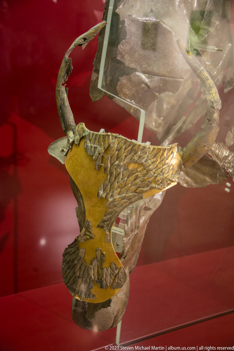 Gold bird-shaped appliqué with spread wings and patterned feathers displayed inside a reflective museum case.