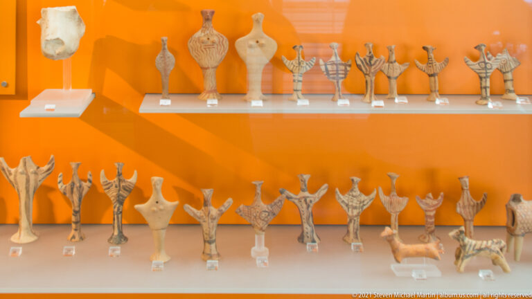 Museum case with rows of small ancient human and animal votive figurines against an orange background.