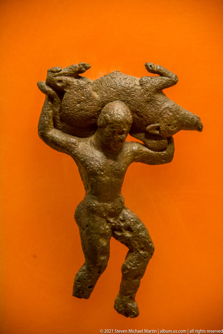 Small bronze figure of a crouching man carrying a large four-legged animal across his shoulders.