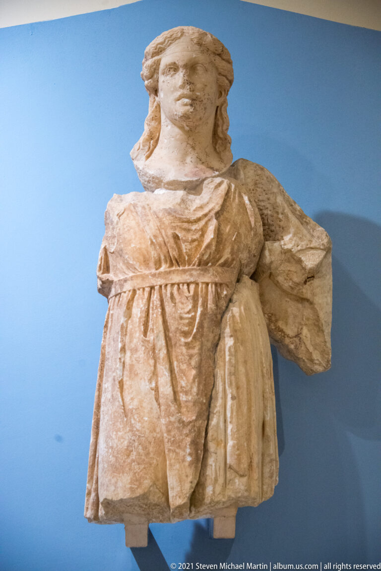 Fragmentary marble female statue with belted drapery, modeled face and wavy hair displayed against a blue wall.