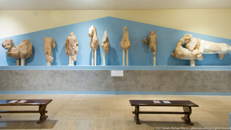 Row of fragmentary ancient sculptures on supports against a blue museum wall, with benches in the foreground.