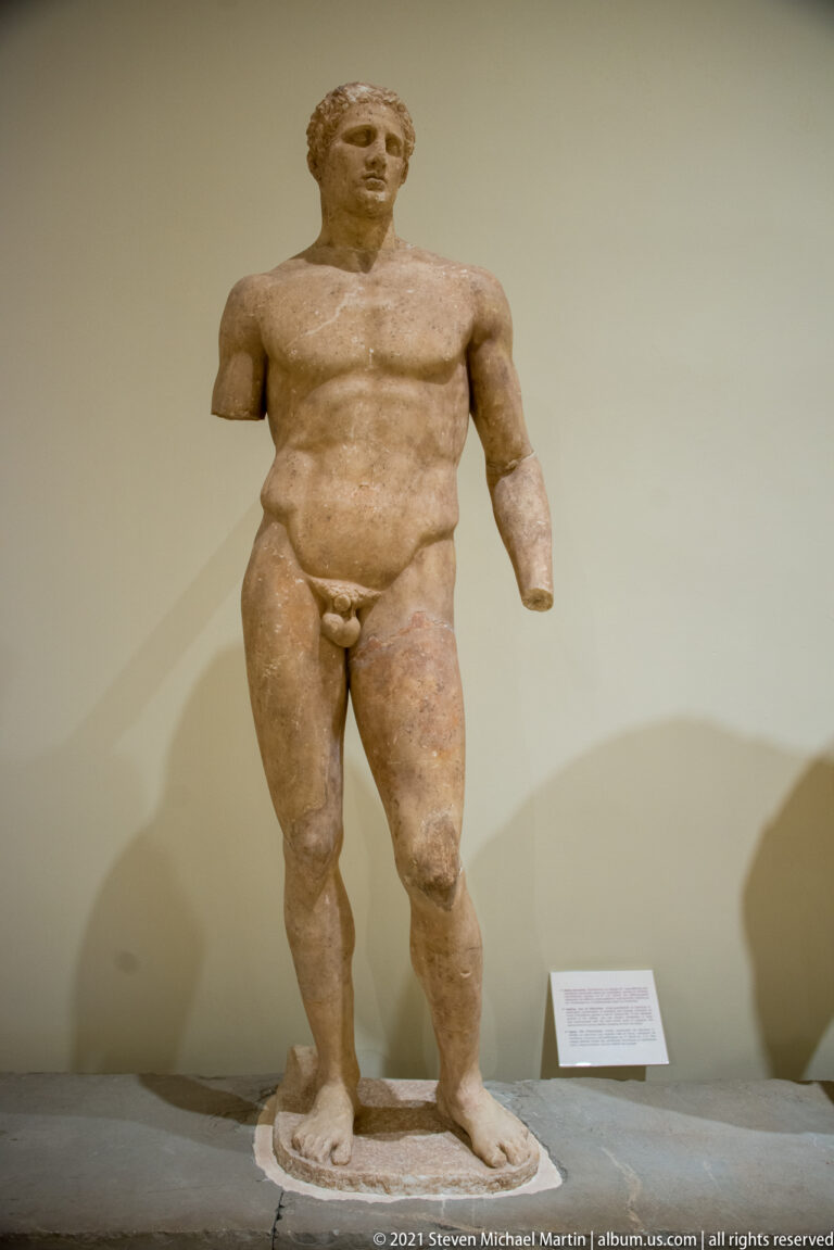 Standing marble nude male statue with intact head and torso but missing both forearms.