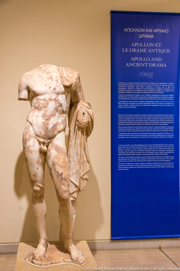Headless marble male statue with drapery over one arm beside a blue panel about ancient drama.