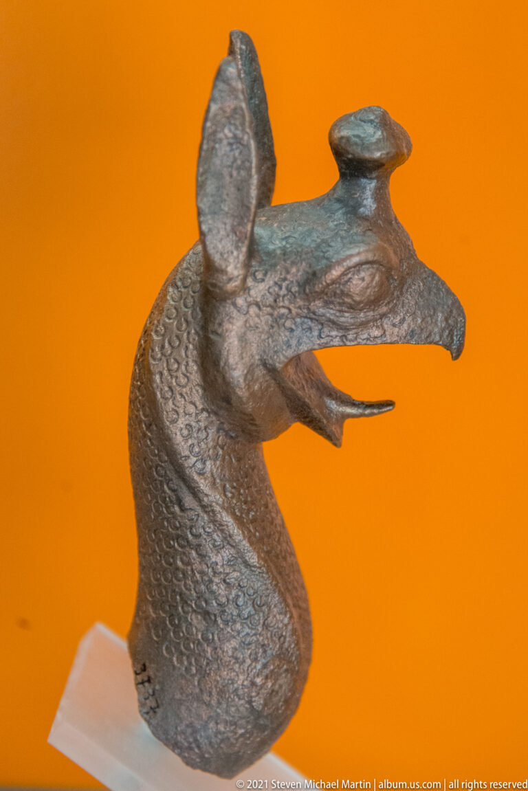 Bronze fantastic animal-head attachment with long muzzle, upright ears, curved neck, and green-brown patina.