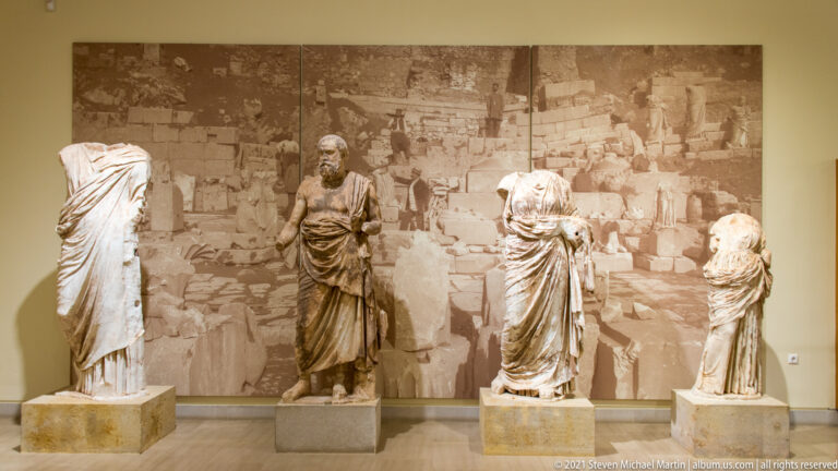 Four draped ancient statues displayed in front of a large sepia excavation photograph.