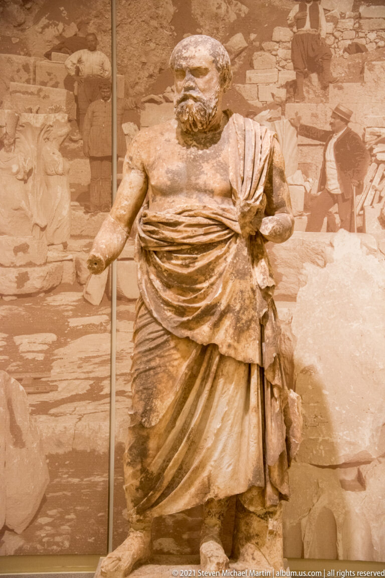 Bearded ancient male statue in a short folded garment, with one arm extended and the other missing.