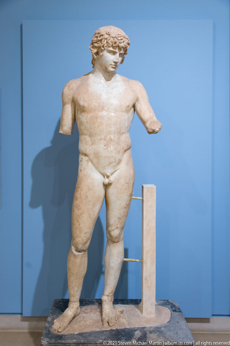 Marble nude youth with thick curled hair, intact legs and torso, and both forearms missing.