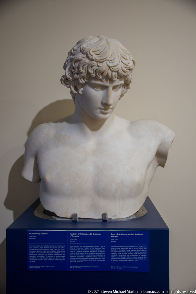 Marble bust of a youthful male with thick curls, downturned face, bare chest, and broken shoulders.