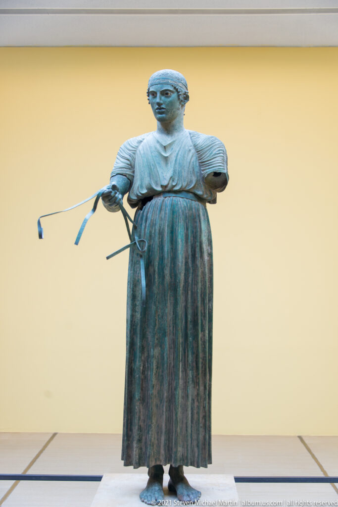 Full-length bronze Charioteer of Delphi in a long pleated garment, holding surviving reins in one hand.