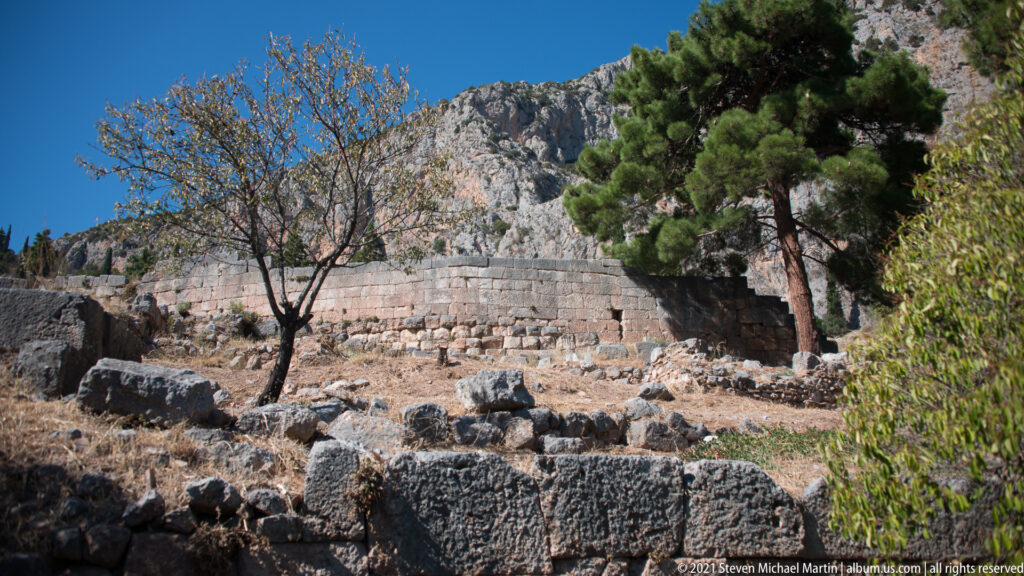 Curving wall of large stone blocks on a rocky, tree-lined slope beneath the cliffs at Delphi.