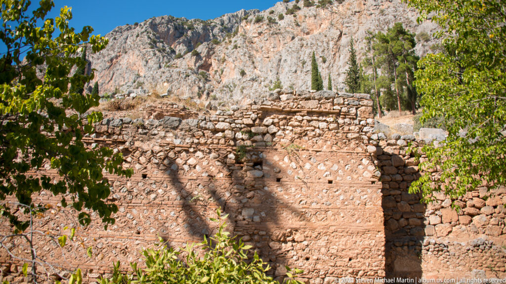 Reddish brick and rubble masonry ruin with a surviving curved or vaulted section among trees at Delphi.