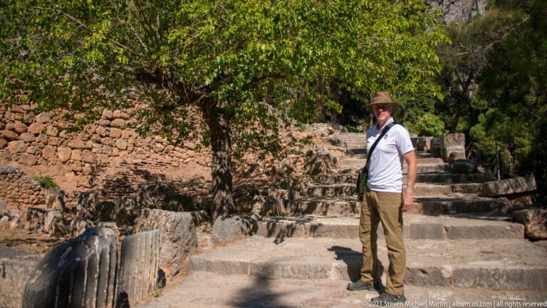 Visitor standing on broad stone steps between low ancient walls and scattered blocks at Delphi.