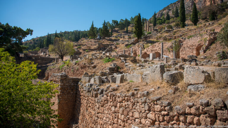 Terraced stone ruins, foundations and column fragments climbing a steep hillside with cypress trees at Delphi.