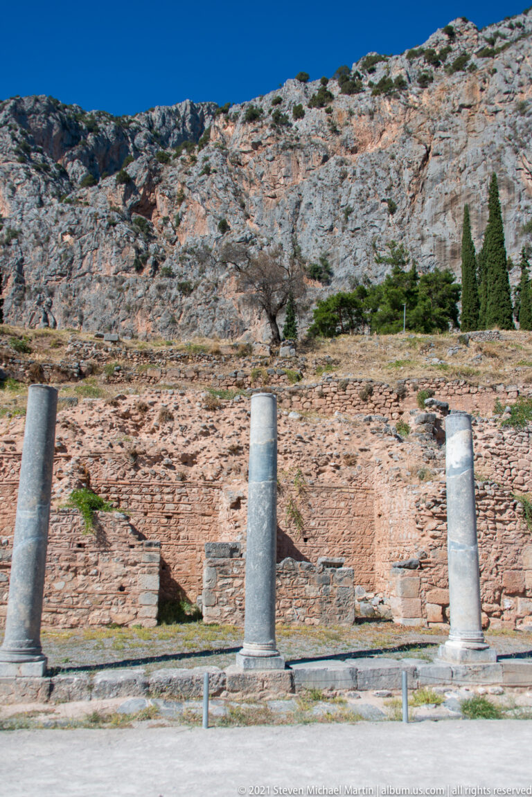 Three pale ancient column shafts aligned before reddish masonry ruins and a rocky mountain slope.