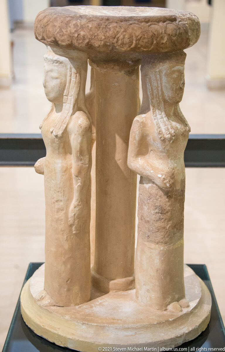 Pale stone support with three carved female figures arranged around a central cylinder beneath a round slab.