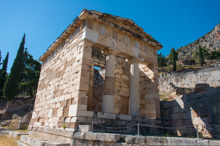 Restored stone Treasury of the Athenians with two Doric columns at the porch against the rocky Delphi hillside.