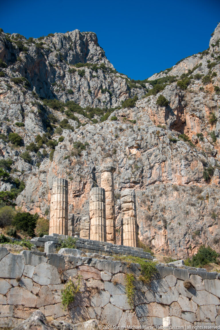 Three ancient Doric columns standing below steep rocky cliffs at the Temple of Apollo terrace in Delphi.