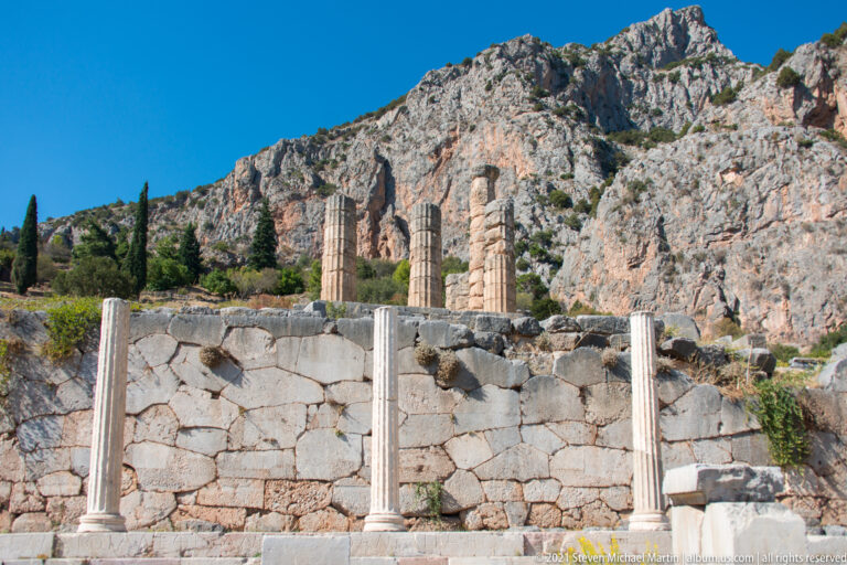 Standing Doric columns, fallen drums, and a stone retaining wall beneath the rocky cliffs at Delphi.