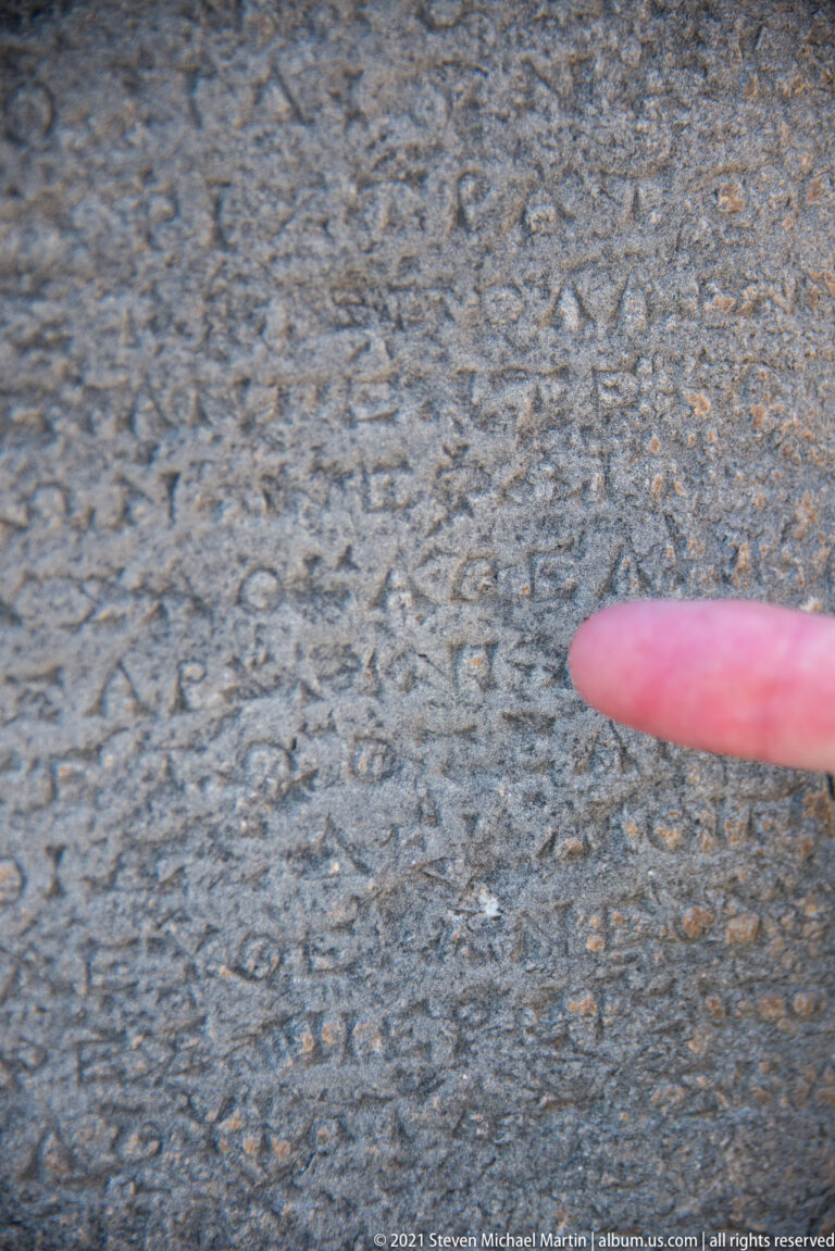 Fingertip pointing at small Greek letters incised across a dark, weathered stone slab.