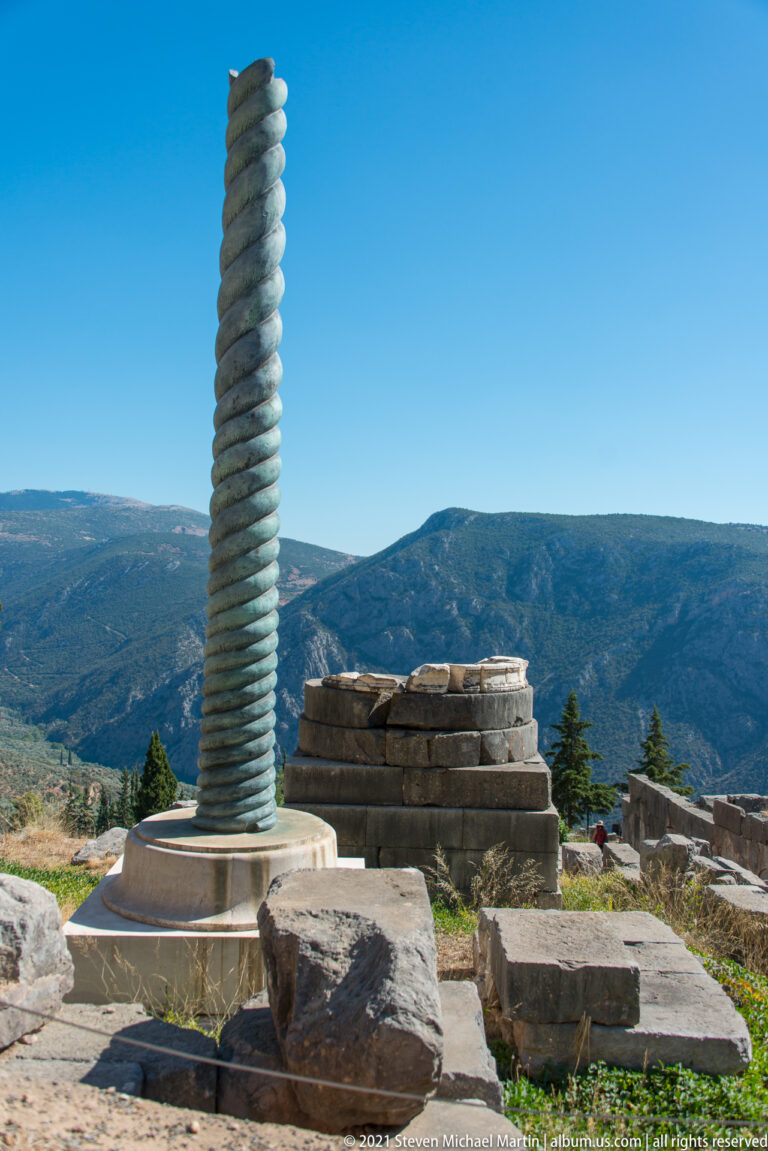 Spiral Serpent Column reconstruction on a stone base, with ruins and broad mountain ridges beyond.