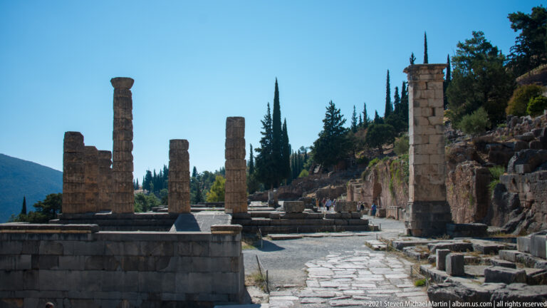 Ancient paved route receding uphill between standing columns, walls, and scattered stone remains at Delphi.