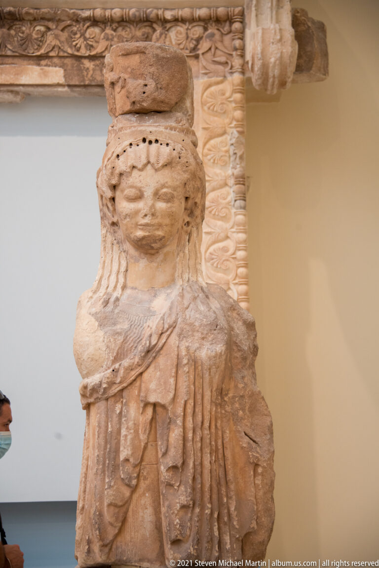 Archaic stone female figure with long hair and layered drapery supporting an ornate carved architectural block.