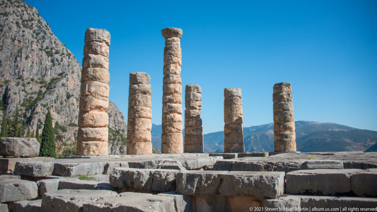 Six standing Doric columns on the ruined Temple of Apollo platform, with mountains and blue sky beyond.