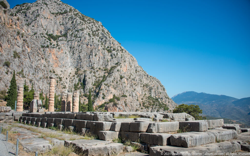 Wide view of the Temple of Apollo platform, standing columns, foundation blocks, and steep cliffs at Delphi.