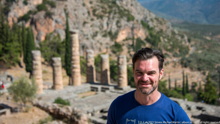 Smiling man in a blue shirt standing in front of blurred Temple of Apollo columns and mountain cliffs.