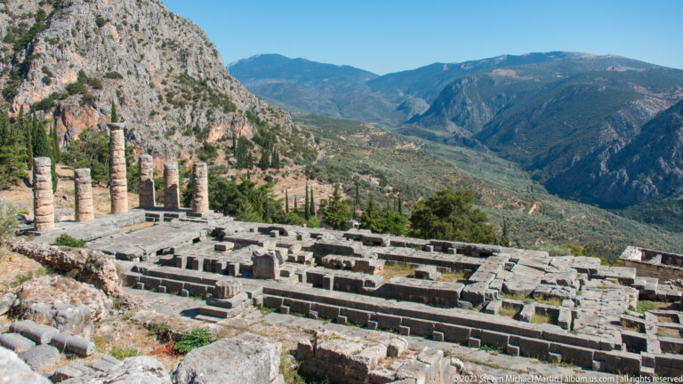 Elevated view over the Temple of Apollo ruins and standing columns toward the broad mountain valley beyond.