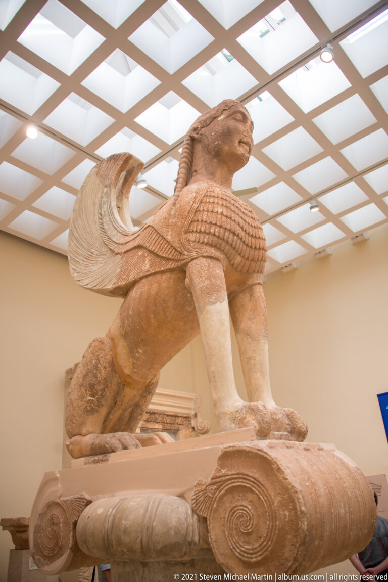 Oblique side view of the Sphinx of Naxos, showing lion body, raised wing, forelegs, and Ionic capital.