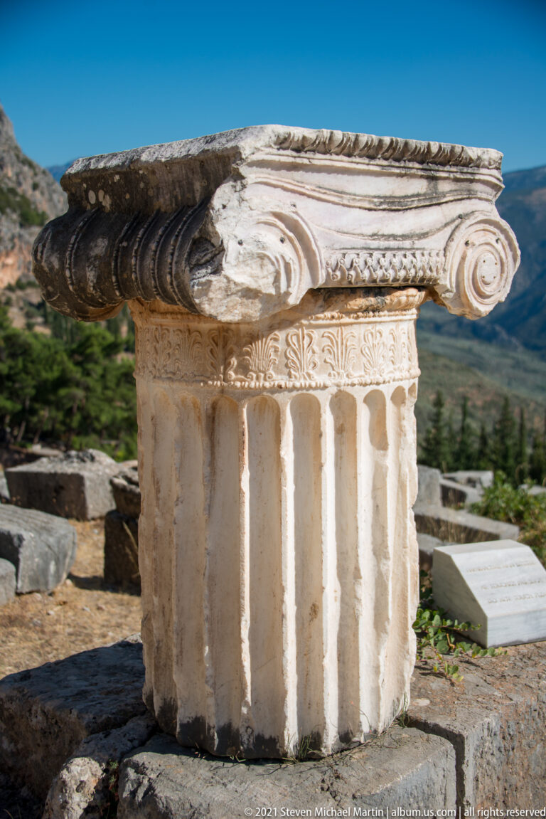 Large carved Ionic capital with spiral volutes and a deeply fluted stone shaft displayed outdoors at Delphi.