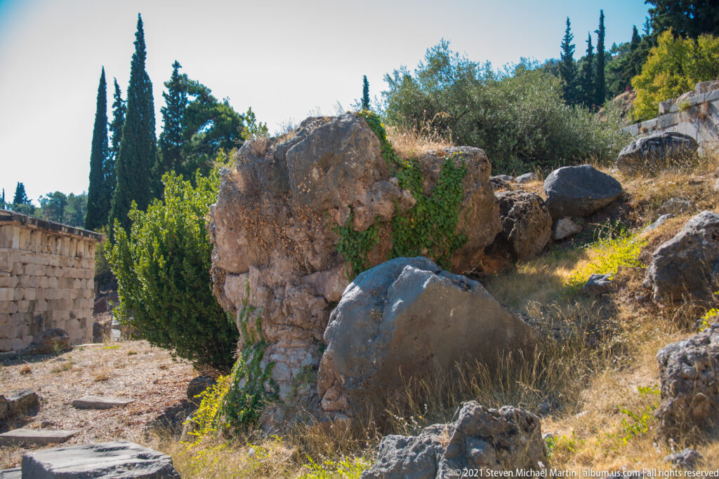 Irregular weathered limestone outcrop surrounded by shrubs, dry grass, and scattered archaeological stonework at Delphi.