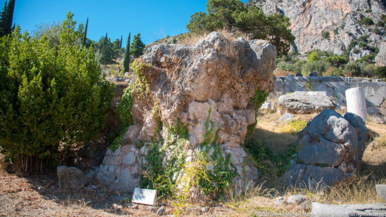 Large irregular limestone formation beside green shrubs, stone fragments, and a small site label at Delphi.