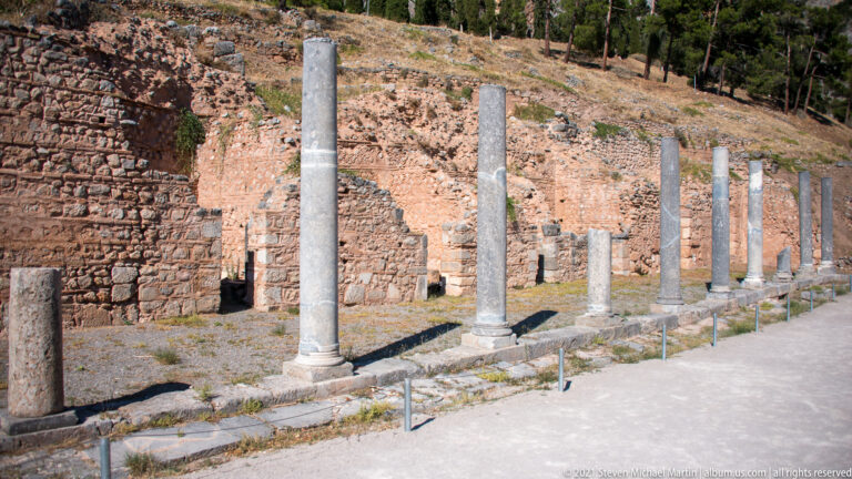 Row of slender standing stone columns beside ancient brick-and-stone walls on a sloping archaeological site.