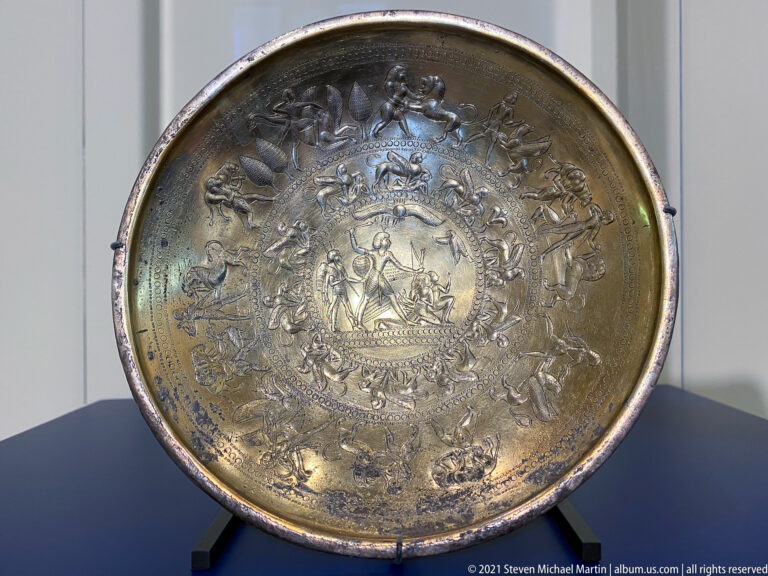 Shallow metal bowl displayed upright, densely embossed with concentric bands of figures, animals, and ornament.