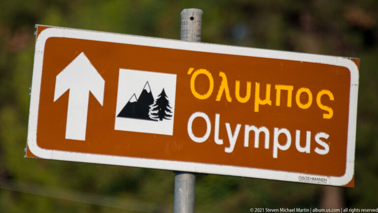 Brown Greek-English road sign with an upward arrow, mountain pictogram, and the words Όλυμπος and Olympus.