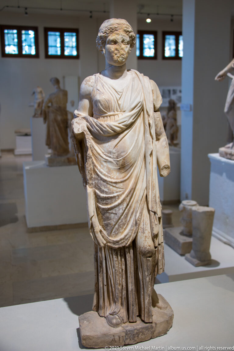 Standing marble man in a heavy wrapped mantle, with one forearm missing and deeply carved folds across the torso.
