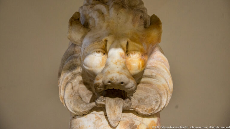 Close view of a pale stone lion head with open mouth, projecting tongue, rounded muzzle, and carved mane.