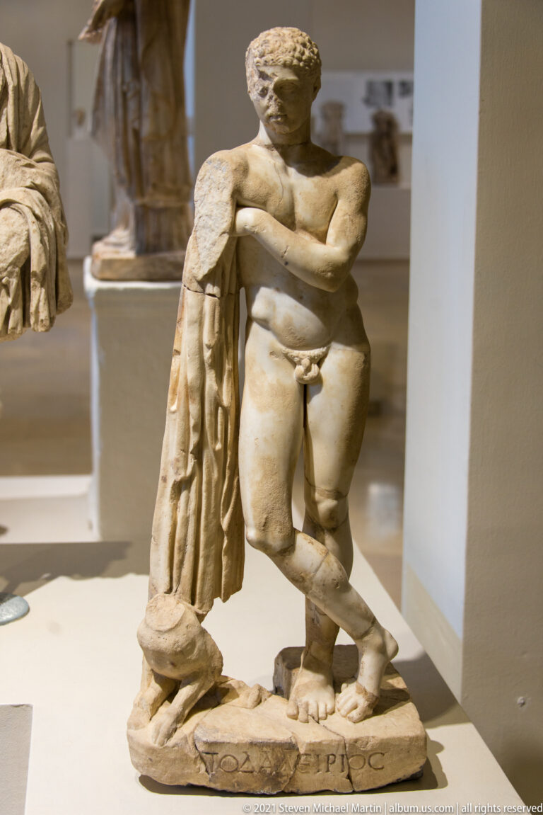 Small nude marble male figure with crossed legs beside a draped vertical support, with one arm and lower limbs incomplete.