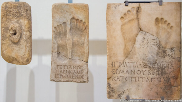 Stone plaques with carved human footprints, visible toes, and Greek inscriptions displayed together on a museum wall.