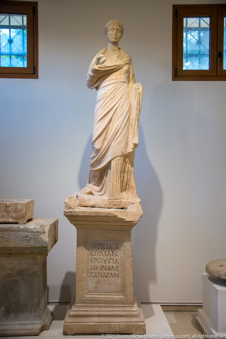 Large seated marble female figure in layered drapery on a tall rectangular pedestal bearing a Greek inscription.